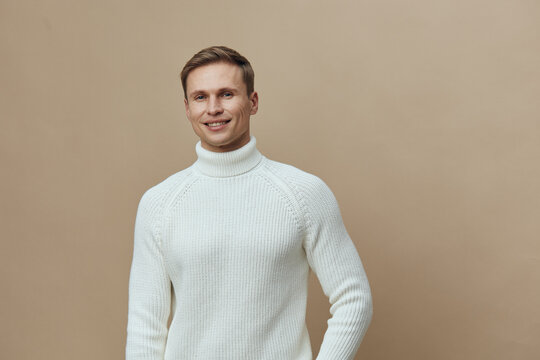 Smiling young man wearing white knitted turtleneck jumper pose on plain beige background studio portrait. People lifestyle concept, casual fashion, happiness, confident, male model, autumn winter