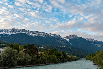 A serene river flows through a lush green valley, framed by towering snow-capped Alpine mountains under a vibrant sky with scattered clouds.