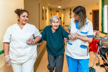 Nurses helping elderly woman walking in nursing home corridor
