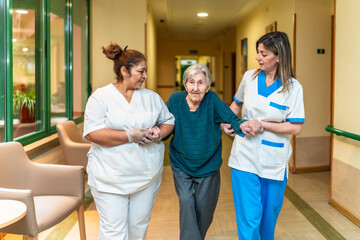 Nurses helping elderly woman walking in nursing home corridor