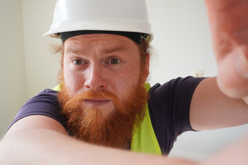 A bearded man in a white hard hat and bright yellow safety vest takes a close-up selfie. His...