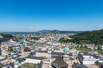 Aerial view of Salzburg, Austria, featuring historic baroque architecture, church domes, and the Salzach River winding through the city, with mountains in the background on a clear summer day.