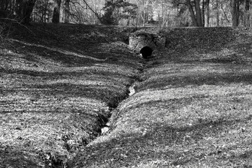 Black and white landscape of a ravine with a small stream and a black drain hole made of masonry with beautiful chiaroscuro