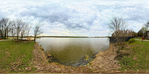 Panoramic 360-degree view of a lakeshore surrounded by early spring trees with sparse leaves. A...