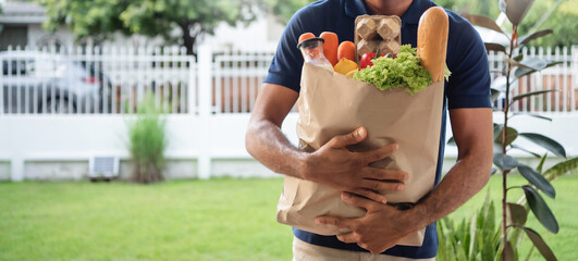 African American delivery man holding paper bag filled with fresh groceries, including vegetables, bread, and carton, stands outdoors in garden setting. scene sense of healthy food © eggeeggjiew