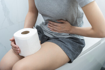 Stomach ache, sick asian young woman holding stomach pain and holding tissue paper roll in bathroom...