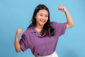 Fototapeta premium Energetic young Asian woman smiling confidently while flexing her arms in a strong pose. Her positive and enthusiastic expression is perfect for advertising, lifestyle, or promotions.