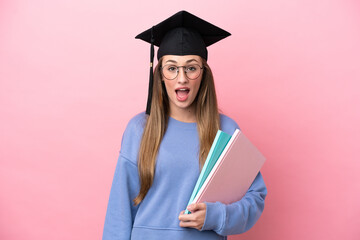 Young student woman wearing a graduate hat isolated on pink background with surprise facial...