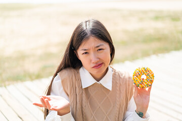 Young Chinese woman holding a donut at outdoors making doubts gesture while lifting the shoulders