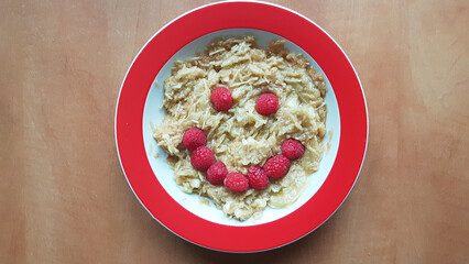 Children's plate with muesli and grated apples. A face made of raspberries. On a wooden table.