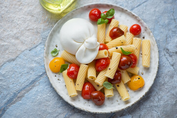 Pasta with burrata cheese and roasted cherry tomatoes, horizontal shot on a blue and beige stone background, high angle view