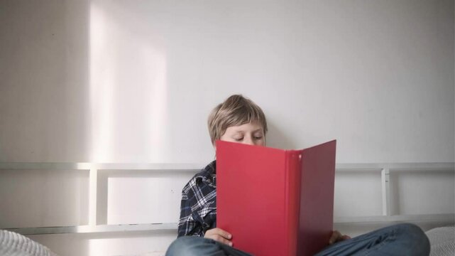 Young boy in plaid shirt sitting on bed, reading carefully from a red folder at cozy home interior