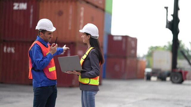 Two logistics workers in safety vests and helmets discussing shipping operations with laptop and radio in a container yard. Coordination, teamwork, supply chain, and transport.