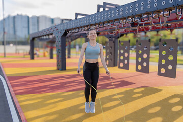 A young woman in sports clothes jumping rope on a fitness playground. She smiles and looks energetic. Exercise equipment and trees are visible in the background.
