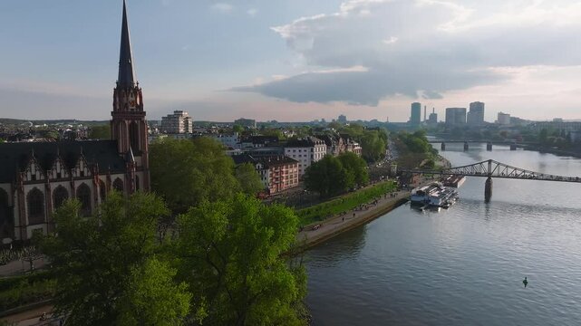 Aerial view of Frankfurt, Germany, showcasing the Main River, modern skyscrapers, Eiserner Steg, docked boats, and a church with a red steeple.