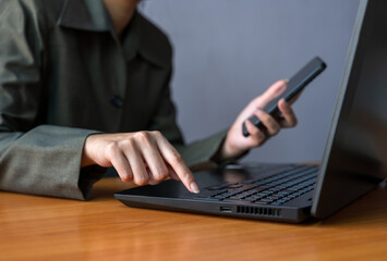 Woman in green blazer multitasking at office wooden desk. Female using laptop with one hand while holding smartphone in the other