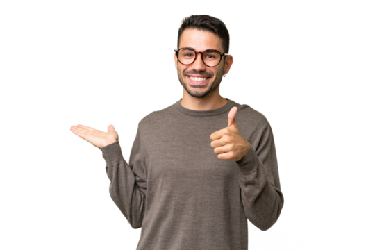 Young handsome caucasian man over isolated background holding copyspace imaginary on the palm to insert an ad and with thumbs up
