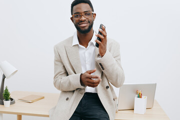 Smiling man with glasses holding smartphone in office setting, dressed in beige blazer and white shirt, standing near desk with minimal decor, modern business lifestyle concept.