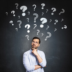 Thoughtful businessman standing in front of a dark wall with floating question marks symbolizing curiosity, problem-solving, and brainstorming.