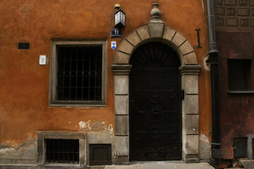Vintage arched black metal door with decorative details in a stone frame, set into an old textured orange wall with weathered surfaces and barred windows — historical European architectural detail