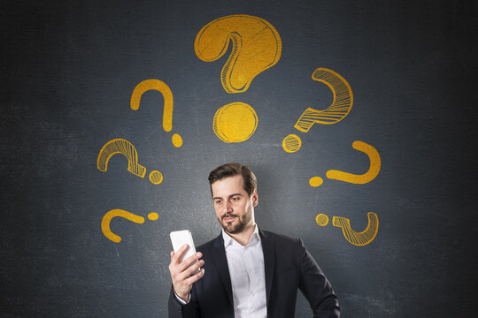 Businessman looking at smartphone surrounded by yellow question marks on dark textured background with abstract light design concept..
