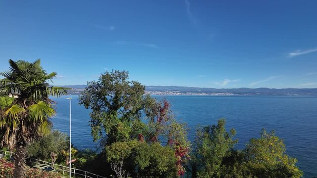 Luxury room with sea view in Lovran, Istria - Croatia. There is a palm tree in the foreground and a mountain range in the background. Seaside balcony on a sunny morning with modern glass railing.