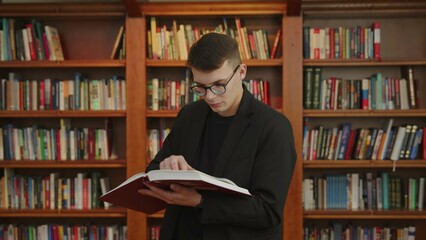 A young man is deeply engrossed in reading a book while seated in a library environment. Media