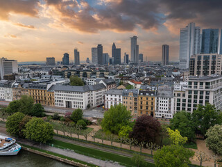 Obraz premium Aerial view of Frankfurt, Germany, featuring modern skyscrapers like Commerzbank Tower, a riverside promenade, a docked boat, and a dramatic sunset sky.