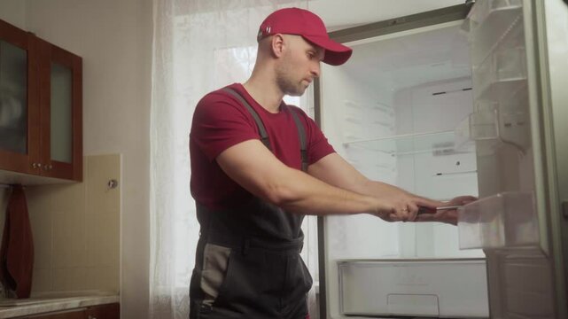 Professional repairman carefully inspects various refrigerator components, including shelves and drawers, during a service call at a residential kitchen
