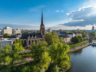 Aerial view of Frankfurt, Germany, featuring Dreikonigskirche near the Main River, surrounded by trees, with a cityscape of modern and traditional buildings.