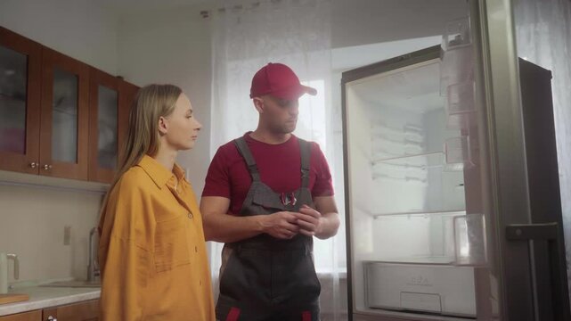 Experienced refrigerator repair technician professionally explaining refrigerator shelving mechanism to housewife inside residential kitchen during maintenance service visit