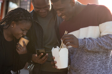Three african american friends laughing and looking at a smartphone while eating street food. Casual and happy atmosphere captured in a candid moment.