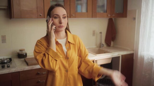 A woman in a yellow shirt stands in her kitchen while discussing urgent plumbing issues with a repair service regarding a leaky sink that needs immediate attention.