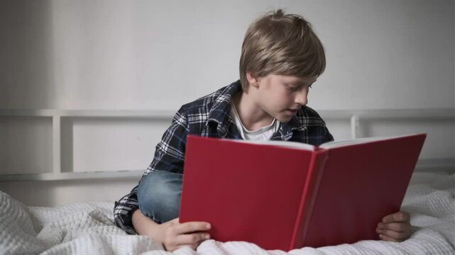 Young boy in plaid shirt sitting on bed, reading carefully from a red folder at cozy home interior