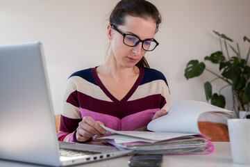 Young woman in glasses looking at papers while sitting at a laptop