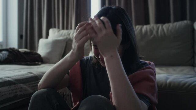 Medium shot of frustrated, distressed 17-year-old Caucasian girl hunched up on floor in living room, scrolling abusive messages on social media, throwing down smartphone and clutching head in despair