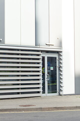 Exterior view of a modern building featuring a minimalist gray and white facade, a glass door with architectural accents, and horizontal blinds.