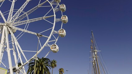 White ferris wheel rotating under blue sky near a sailing ship. Action