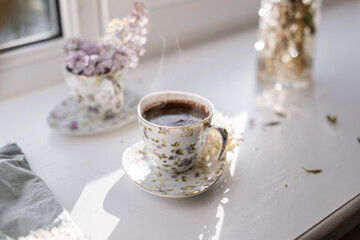 A close-up of freshly brewed coffee in a porcelain teacup, with visible steam and foam, accompanied by a vase of lilac flowers. A serene depiction of springtime comfort and charm.