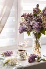 Freshly brewed coffee with foam in a porcelain teacup, accompanied by lilac flowers in a glass vase. Highlights simplicity, elegance, and nature-inspired tranquility.