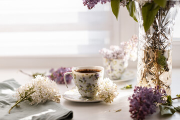 A steaming porcelain teacup of freshly brewed coffee placed beside a lilac bouquet in a glass vase....