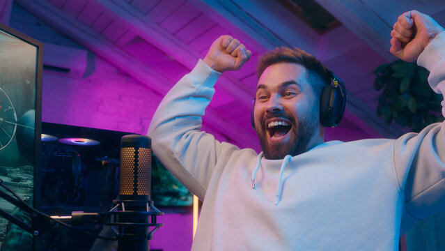 Joyful Caucasian male guy gamer streamer playing computer tech e-sport at home man winning shout winner rejoice addict influencer celebrate game victory champion receive golden cup blogger stream neon