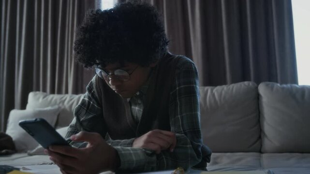 Medium shot of depressed, lonely teenage African American schoolboy, wearing nerdy eyeglasses, sitting alone at home, looking at abusive, violent content on social media, unable to do homework