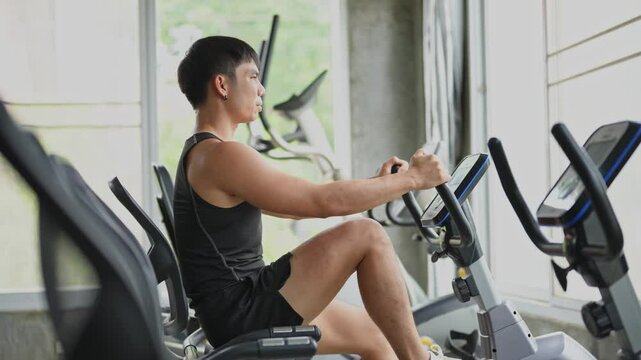 Young Asian man exercising indoors on rowing machine in modern gym, focusing on fitness and strength training while maintaining proper form and posture