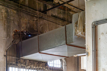 fragment of an abandoned industrial building. The photo shows a rusty and damaged ventilation system suspended on chains from the ceiling. The walls are damaged, with visible traces of moisture, splin