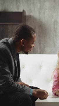 Little girl pupil answers nodding African-American tutor while classmate sits on sofa at home English lesson with native speaker slow motion