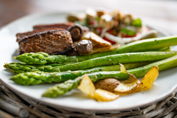 Roasted green asparagus with a beef steak, mushrooms, and crispy potato slices served on a white...