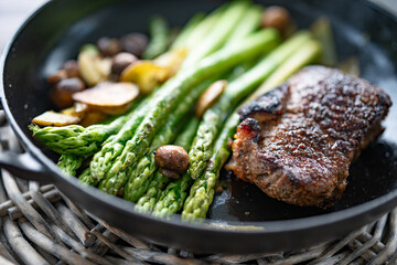 Crispy roasted beef steak with green asparagus, mushrooms, and potato slices in a cast iron pan. Close-up with shallow depth of field.