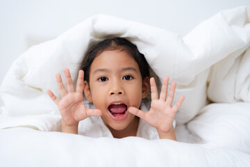 Happy little girl playing hide and seek under blanket in bed