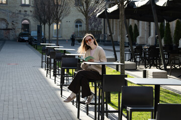 In a tranquil outdoor setting, a woman enjoys reading a book at a cafe table, surrounded by empty chairs and young trees under clear blue skies. It's a peaceful afternoon